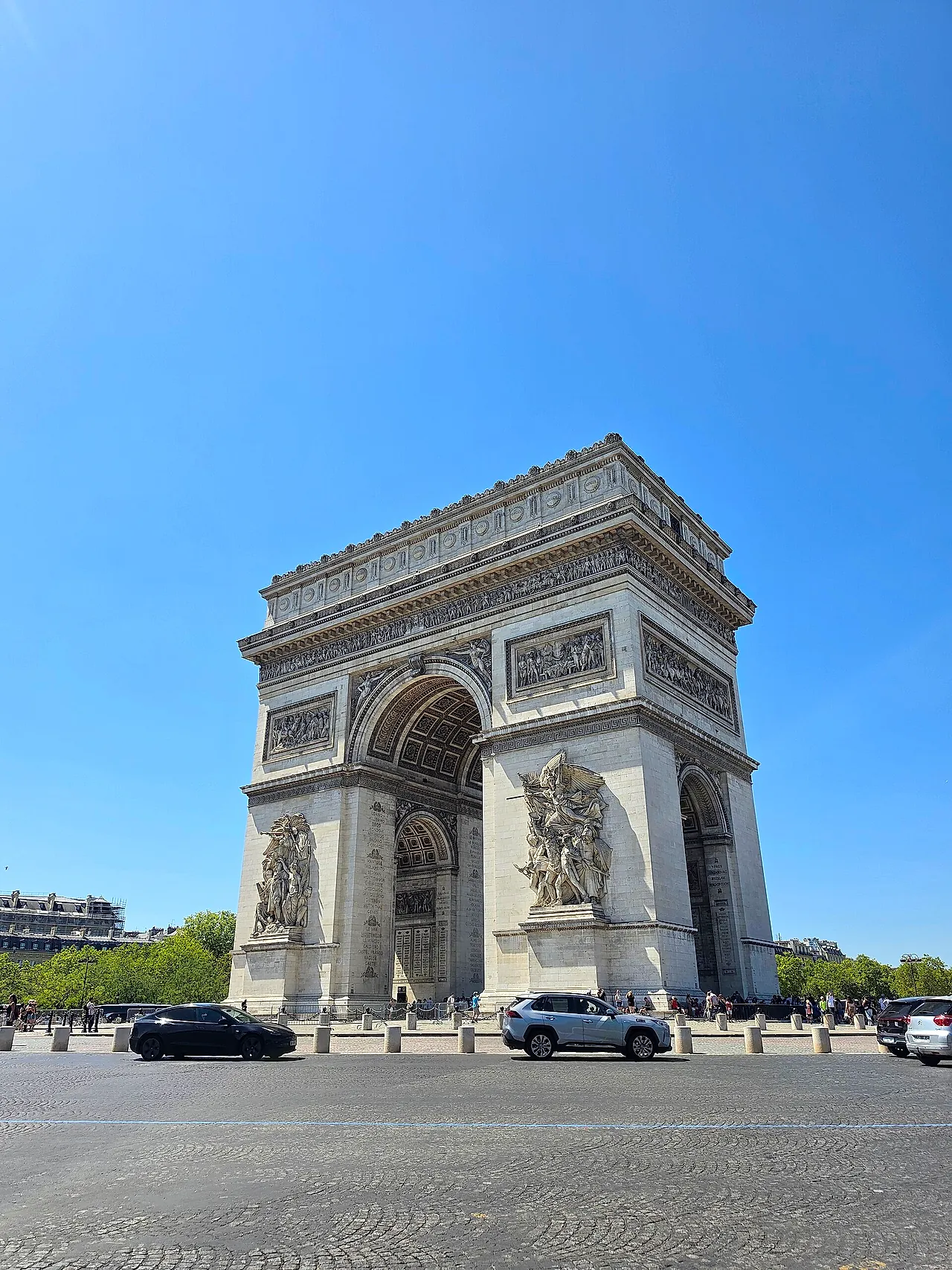 The Arc de Triomphe in the morning light, seen from a corner with its sculpted reliefs and the central arch under a clear blue sky.