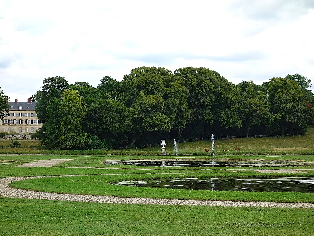 The gardens of the Château de Chantilly: a wide lawn with a path, small jets of water in the pools and a dense line of trees behind.