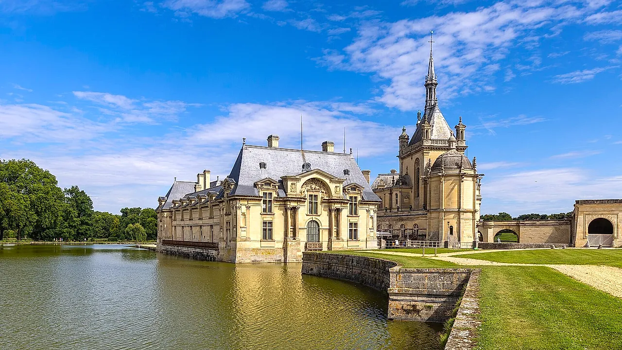 The Château de Chantilly standing at the edge of its water, with a slender tower and slate roofs under a blue sky with light clouds.