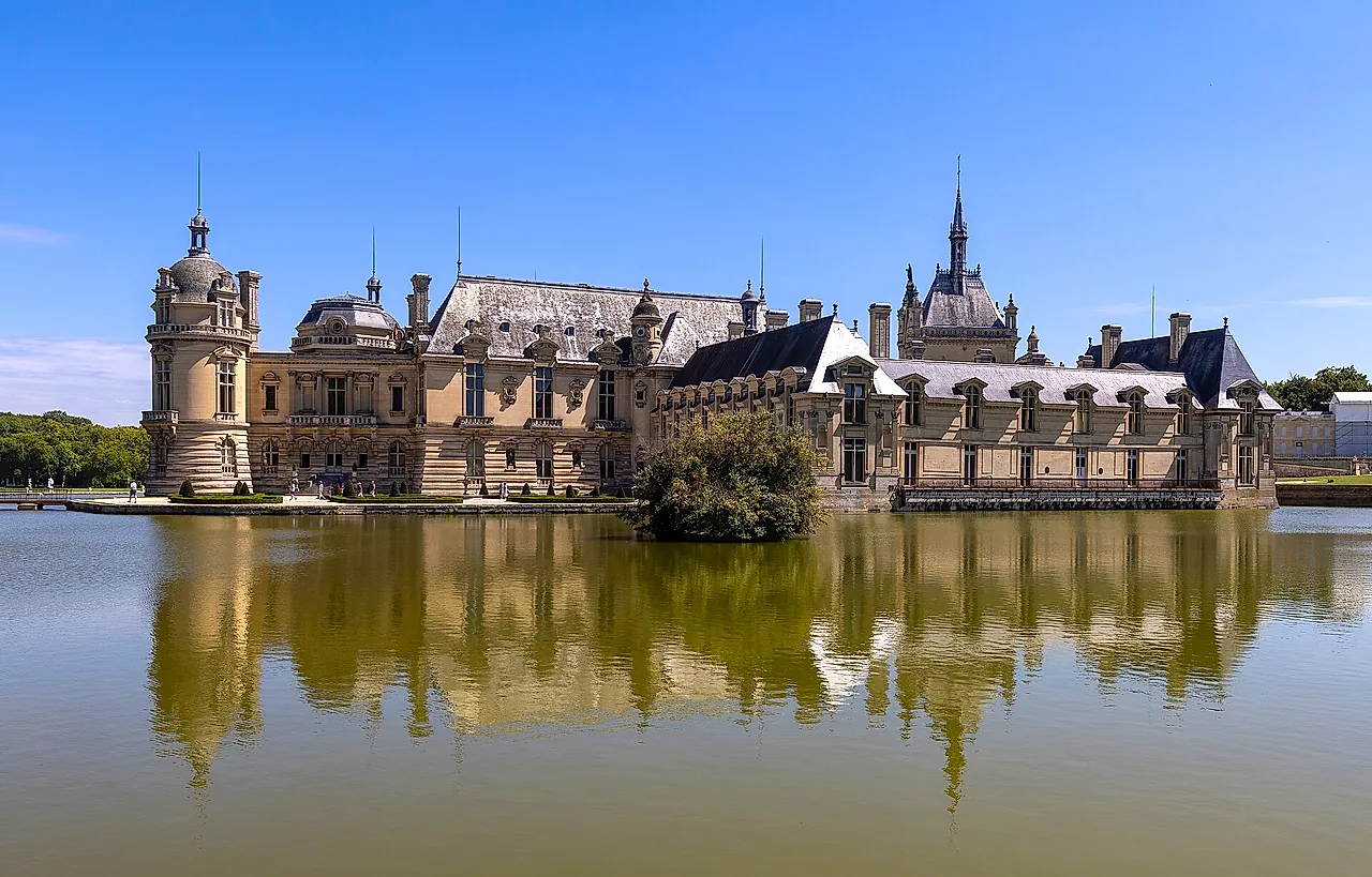 The Château de Chantilly mirrored in the still water of its moat, seen from the paved road on a sunny day.