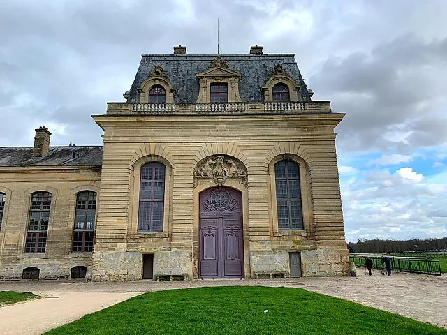 The monumental stone front of the Grandes Écuries at Chantilly, with arched windows and a carved central pediment, above a green lawn.
