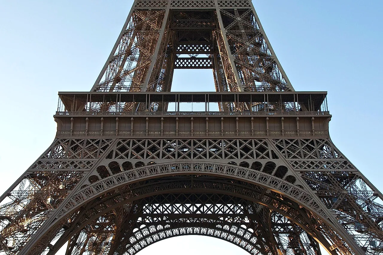 The lower part of the Eiffel Tower seen from the Champ de Mars, with the great arch and the first-floor platform against a pale blue sky.