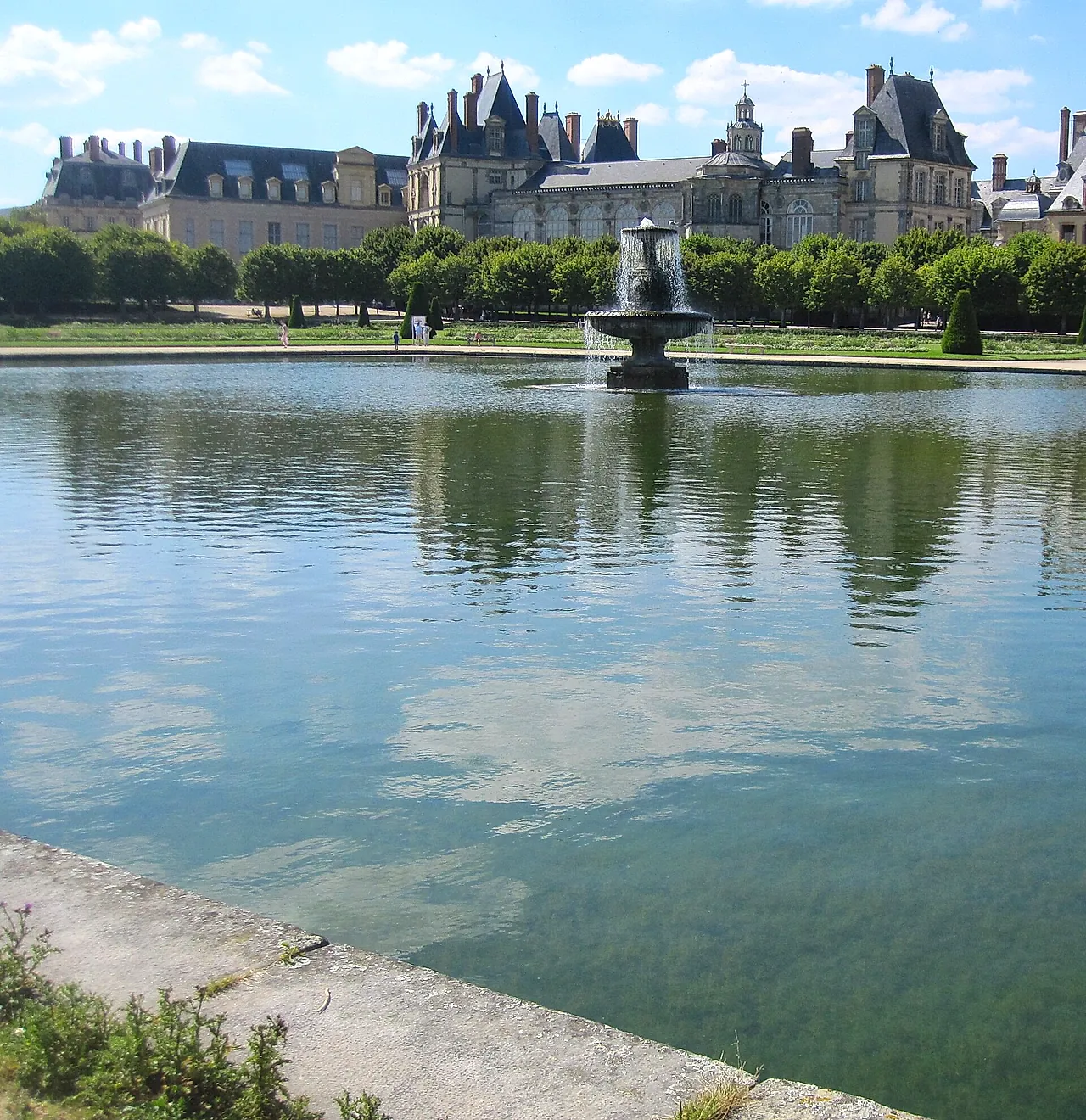 A large still pond in the gardens of Fontainebleau with a fountain in the distance and trees reflected in the water.