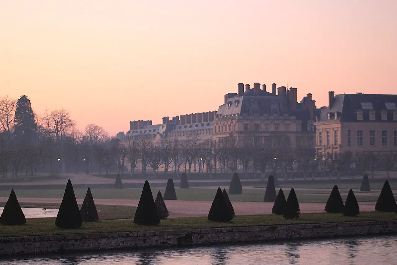 The Château de Fontainebleau at dawn in pink light, behind the trimmed cone-shaped yews of the Grand Parterre.