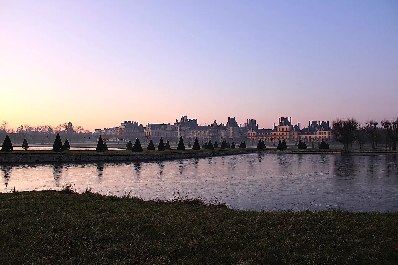 A wide view of the Château de Fontainebleau across the water at dusk, with the Grand Parterre in the foreground.