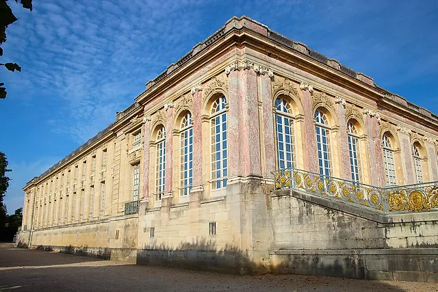 The pink marble and pale stone façade of the Grand Trianon at Versailles, with tall arched windows and a gilded balustrade, against a blue sky.