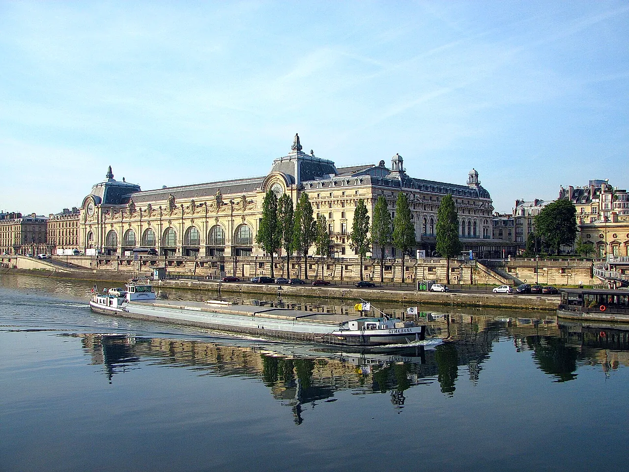 The Musée d'Orsay on the left bank of the Seine, with a barge on the water and its reflection in the river on a clear morning.