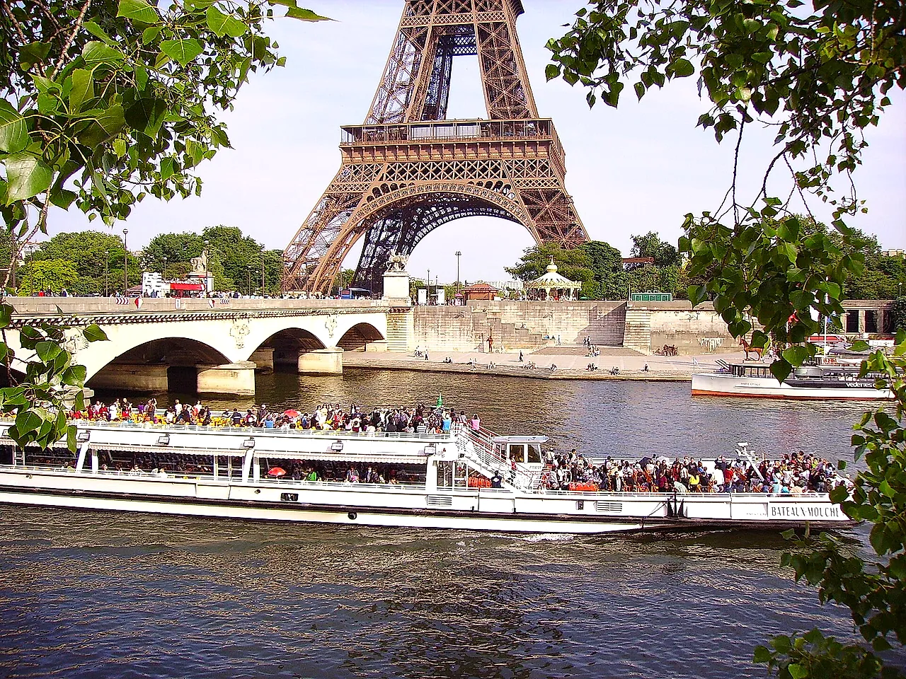 Un battello turistico pieno di passeggeri sulla Senna, con un ponte di pietra e la cima della Torre Eiffel sullo sfondo, tra gli alberi.