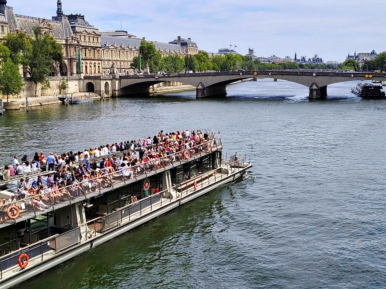 Un battello turistico scoperto e affollato che naviga sulla Senna verso il Pont du Carrousel, con i palazzi della riva sinistra dietro.