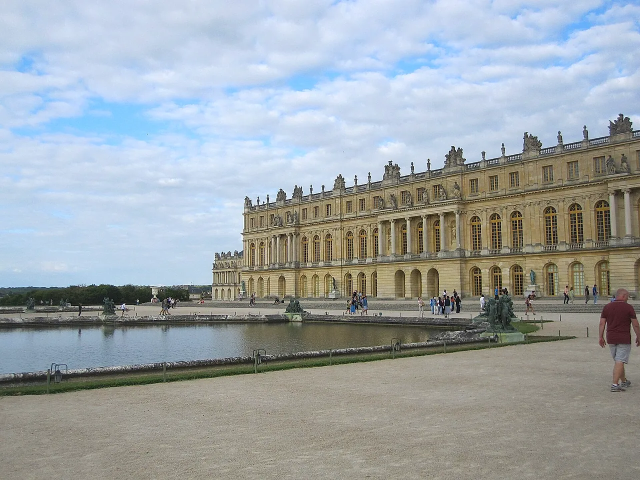 The garden façade of the Palace of Versailles reflected in a large pool, with a few visitors walking along the gravel path in front.