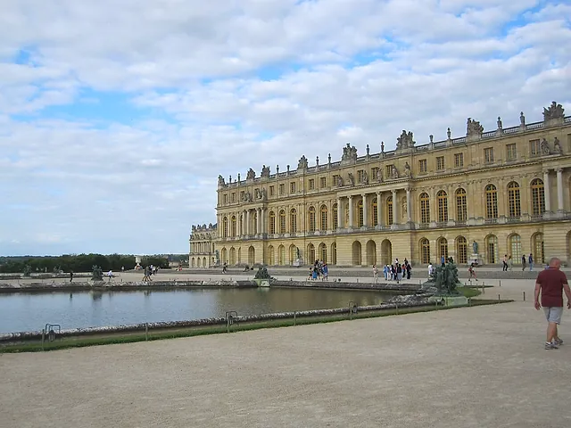 The garden façade of the Palace of Versailles reflected in a large pool, with a few visitors walking along the gravel path in front.