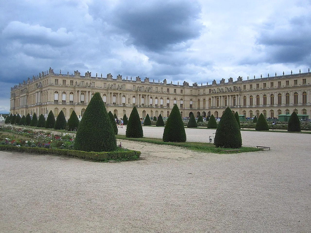 The long façade of the Palace of Versailles seen across the clipped hedges and gravel paths of the gardens, under a cloudy sky.